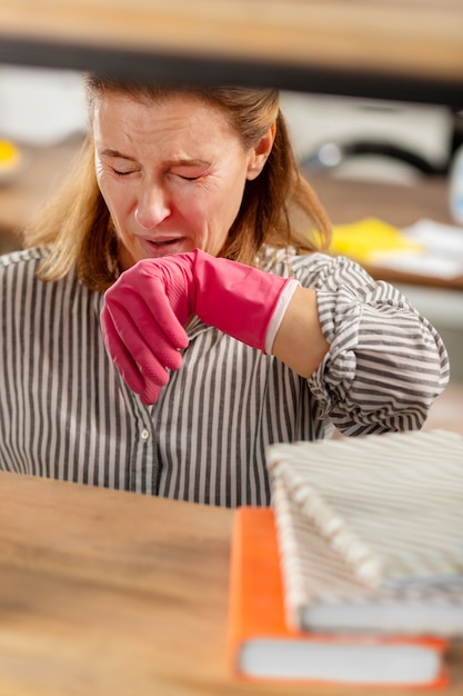 Mature blonde haired woman with facial wrinkles sneezing after dusting shelves