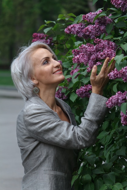 Mature blonde woman in gray suit walks in garden, admires flowers and smiles, selective focus