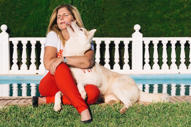 Mature blonde woman seated on the grass with siberian husky dog licking face