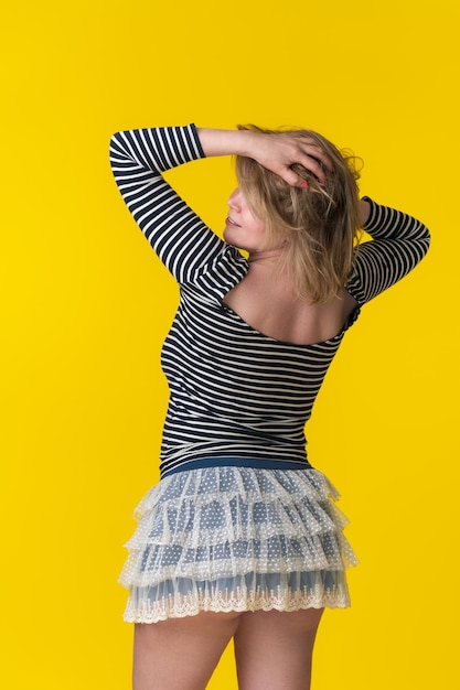Mature blonde woman in striped tshirt and mini skirt standing with hands behind head obscured face