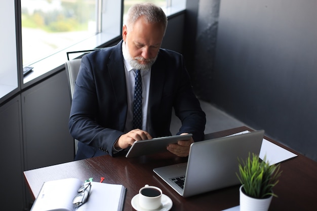 Mature business man in full suit using digital tablet while sitting in the office.