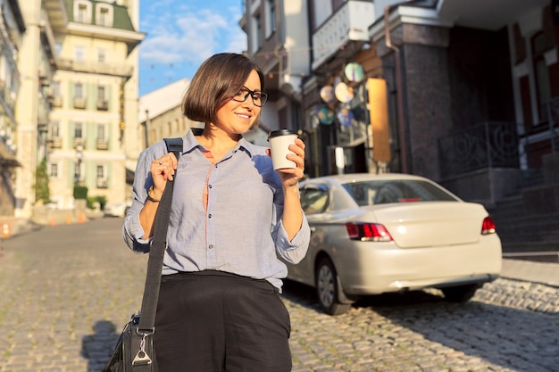 Mature business woman in glasses with office laptop bag walking along city street Happy smiling middle aged female with cup of coffee urban style background