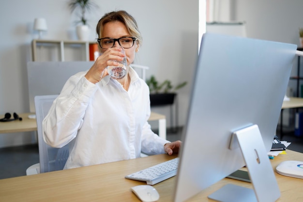 Фото Зрелая деловая женщина в офисе пьет воду во время работы за компьютером