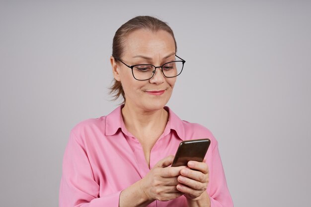 mature business woman with glasses, in a pink blouse, uses a mobile phone, typing a message. isolated photo on a gray background.