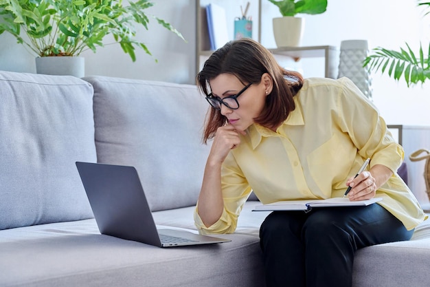 Mature business woman working on couch using laptop making notes in notebook