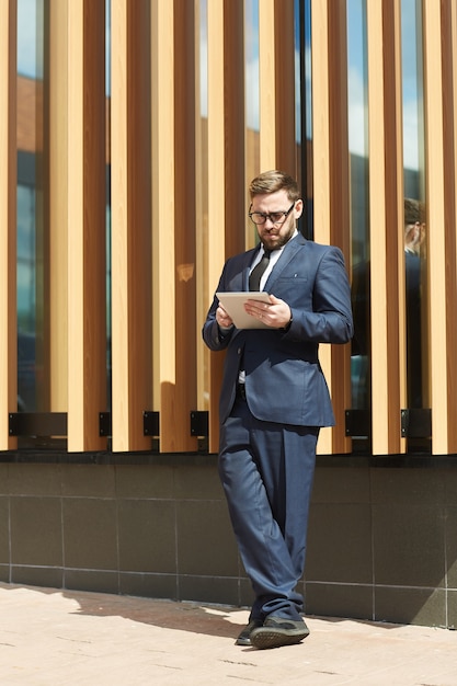 Mature businessman in blue formal suit standing outdoors near the office building and using tablet pc