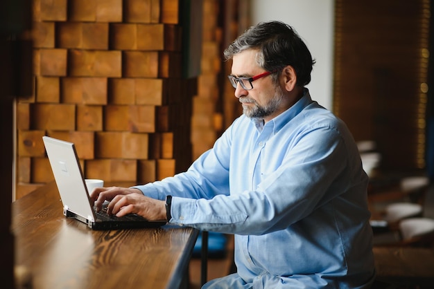 Mature businessman drinking coffee in cafe Portrait of handsome man wearing stylish eyeglasses using laptop looking at camera smiling Coffee break concept