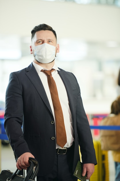 Mature businessman in elegant suit and protective mask standing in lounge of contemporary airport while waiting for flight