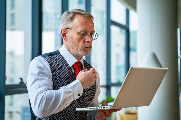 Mature businessman looking at laptop reading and printing\
improvements sitting near the window in his office panoramic city\
view background business concept