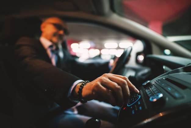 Mature businessman in suit is adjusting a volume on his stereo while driving a car.