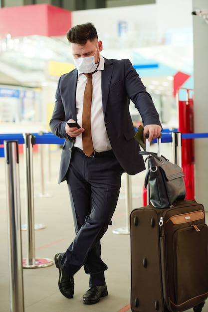 空港で荷物を持って立って、電話でメッセージを入力するスーツを着た成熟したビジネスマン