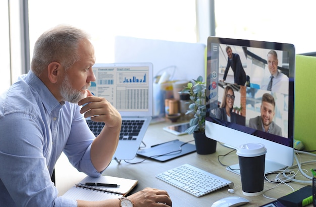 Photo mature businessman talking to his colleagues in video conference. multiethnic business team working from office using computer pc, discussing financial report of their company