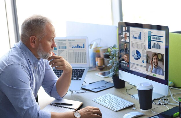 Uomo d'affari maturo che parla con i suoi colleghi in videoconferenza. team aziendale multietnico che lavora dall'ufficio utilizzando un pc, discutendo il rapporto finanziario della propria azienda