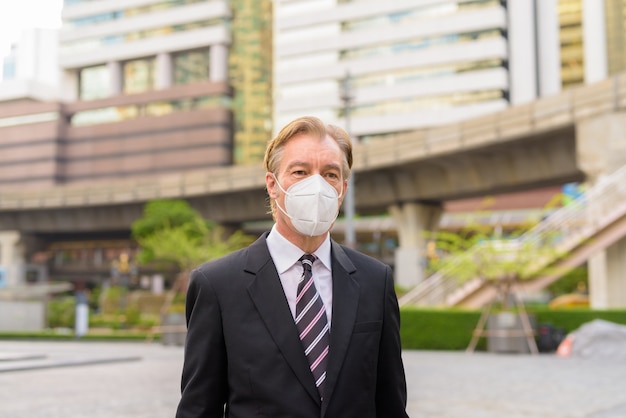 Mature businessman thinking with mask for protection from coronavirus outbreak in the city outdoors