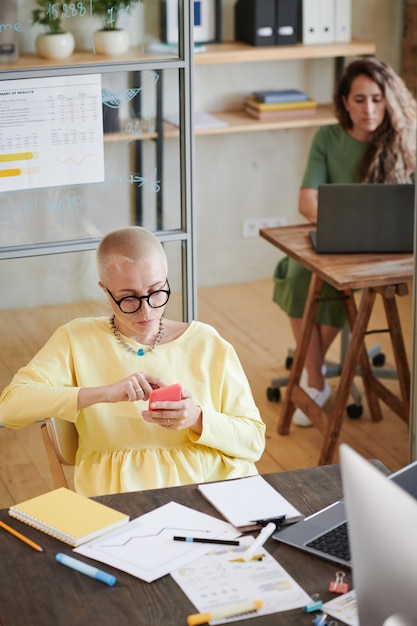 Mature businesswoman in eyeglasses sitting at the table and typing a message on her mobile phone she working online