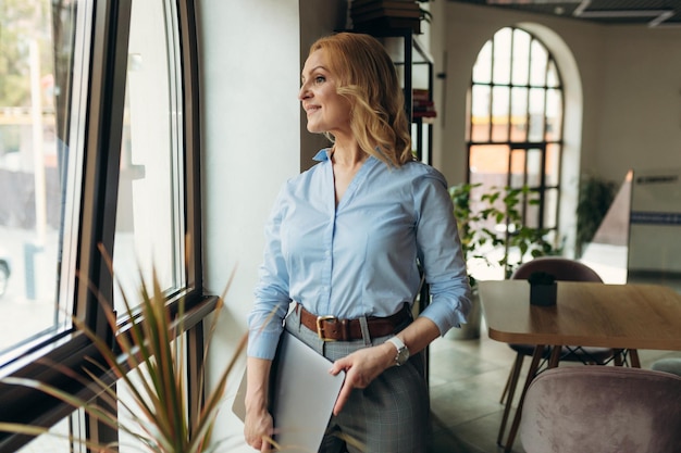 Mature businesswoman holding laptop standing in cafe seen from window