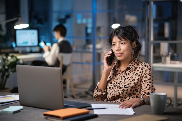 Mature businesswoman sitting at her workplace with documents looking at laptop screen and talking on the phone while working at office in the evening