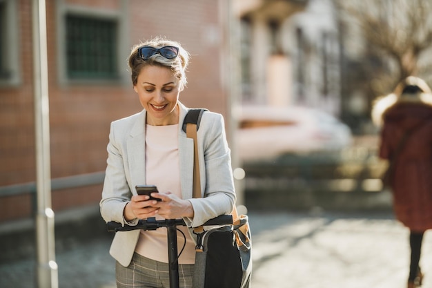 Una donna d'affari matura che usa uno smartphone mentre viaggia con uno scooter elettrico attraverso la città.