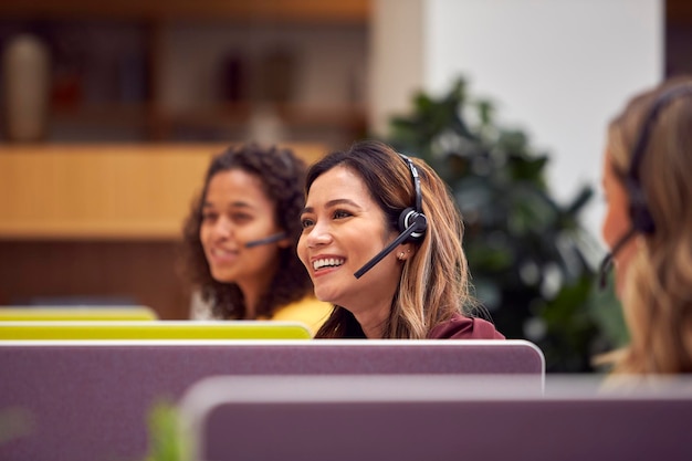 Mature Businesswoman Wearing Phone Headset Talking To Caller In Busy Customer Services Centre