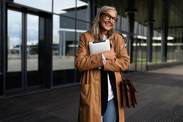 Mature businesswoman with a laptop in her hands against the\
backdrop of the facade of an office