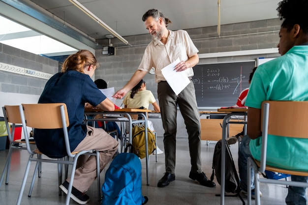 Mature caucasian man teacher hands out sheets of paper to multiracial high school students. Education concept.