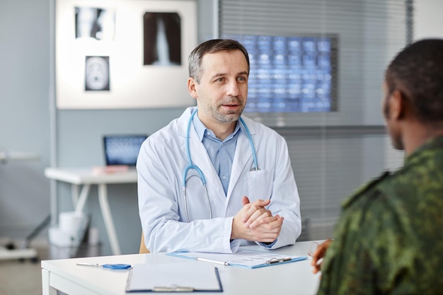 Mature caucasian physician sitting at desk in front of unrecognizable black soldier speaking about t