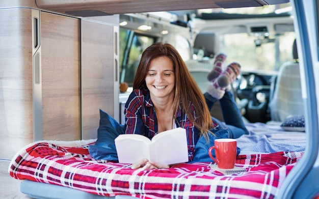 Mature caucasian woman relax inside mini van camper while reading a book and drinking coffee