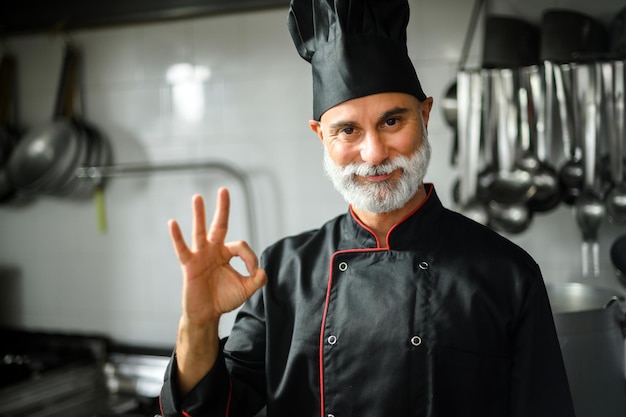 Mature chef in black coat doing the ok sign in his kitchen