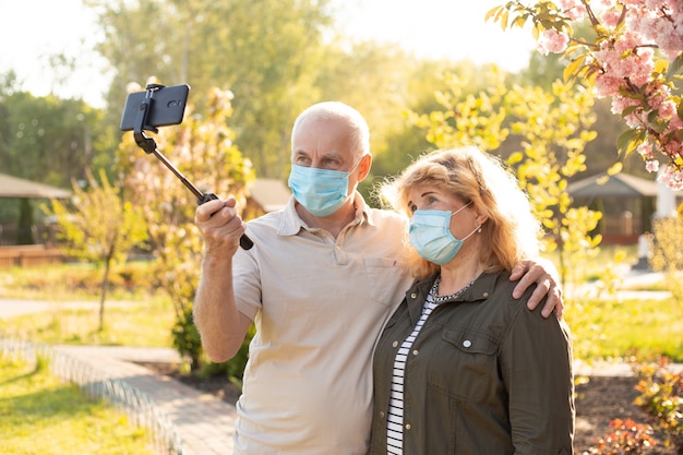 Mature couple making selfie and embracing in spring or summer park wearing medical mask to protect from coronavirus
