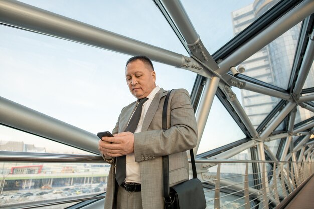 Mature delegate or businessman in formalwear using smartphone while standing in front of camera inside contemporary business center