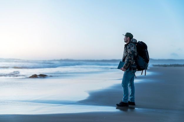 Зрелый цифровой кочевник стоит на берегу со своим ноутбуком и рюкзаком на закате