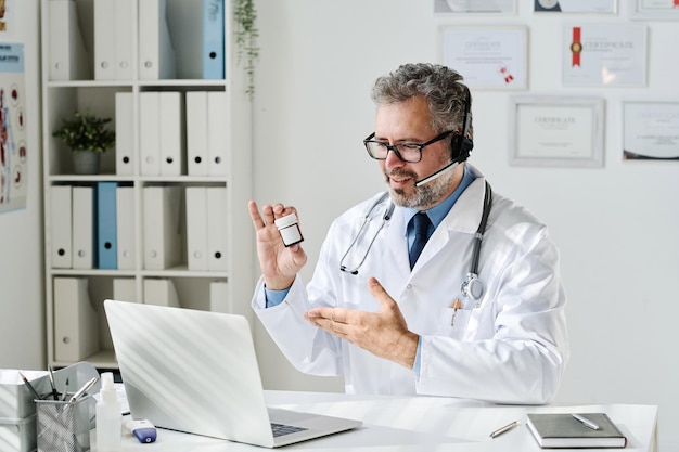 Mature doctor in headphones sitting at his table with laptop\
and prescribing medicine to patient dur