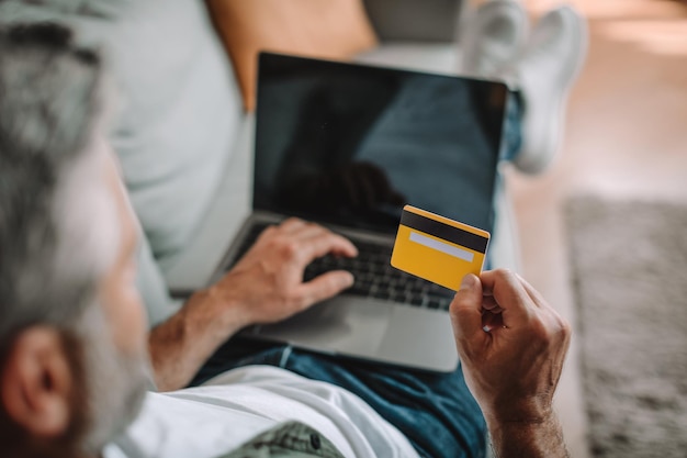 Mature european male resting on sofa and shopping online with credit card and laptop with blank screen