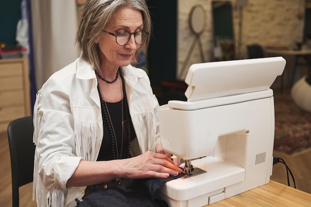 Mature European woman fashion designer tailor sitting at wooden desk using sewing machine sews clothes in a tailoring atelier