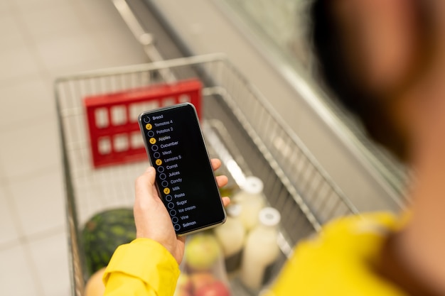 Photo mature female buyer in casual pullover scrolling through shopping list in smartphone while pushing cart with paperbags in supermarket