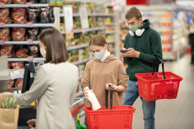 Mature female consumer choosing bottle of champagne while her\
husband with shopping cart going to buy cognac in supermarket