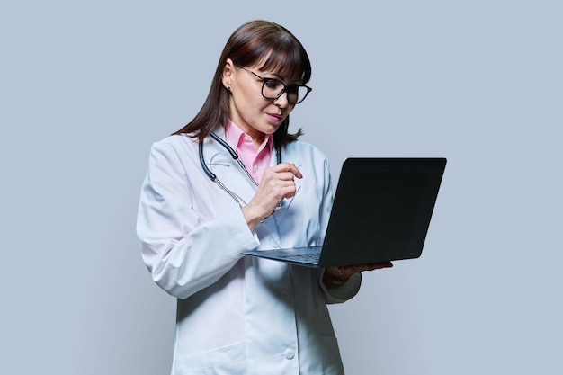 Зрелая женщина-доктор использует ноутбук на сером фоне