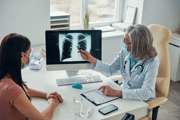 Photo mature female doctor in white lab coat showing to young woman her lungs scan while sitting in her office