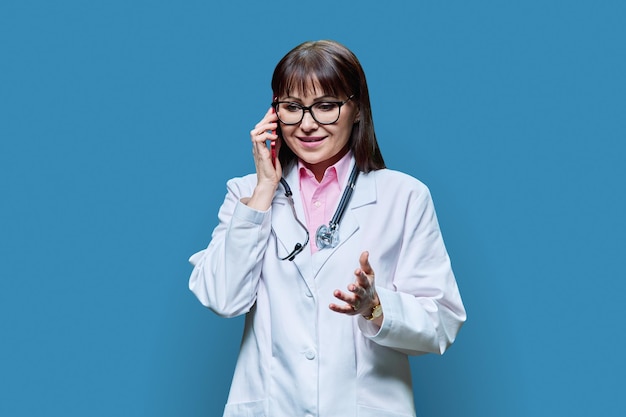 Photo mature female doctor in white lab coat talking on phone on blue background