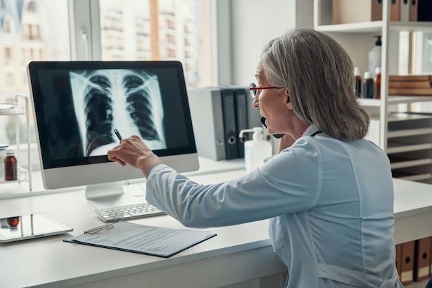 Mature female doctor in white lab coat talking on the phone and examining lungs using computer while sitting in her office