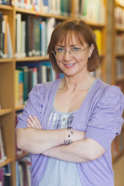 Bibliotecario femminile maturo che posa nella biblioteca con le armi attraversate che esaminano macchina fotografica