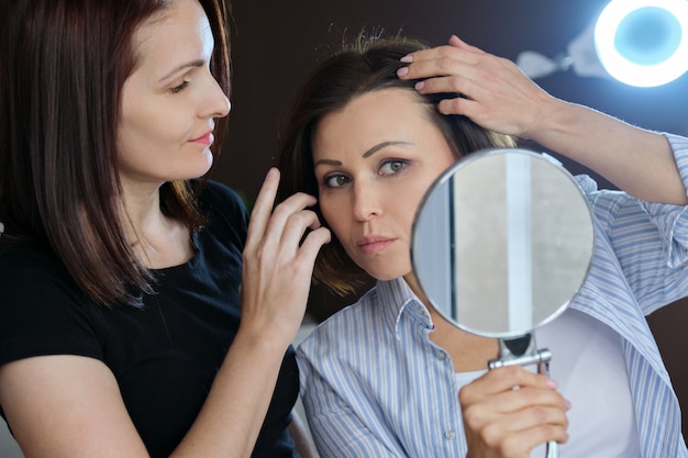 Mature female patient looking in the mirror and female beautician showing on face