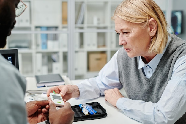 Mature female patient looking at screen of glucometer held by doctor