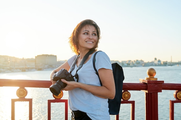 Mature female photographer with professional camera and backpack, smiling female on the bridge on sunny sunset summer day. River, sky, city skyline background