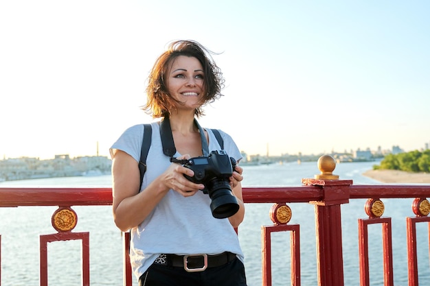 Mature female photographer with professional camera and backpack, smiling female on the bridge on sunny sunset summer day. River, sky, city skyline background