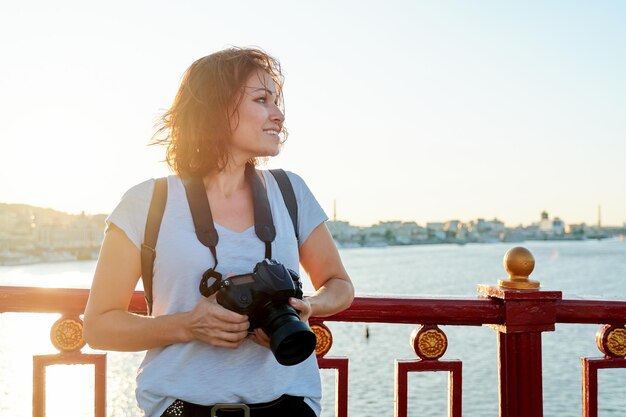 Mature female photographer with professional camera and backpack, smiling female on the bridge on sunny sunset summer day. River, sky, city skyline background