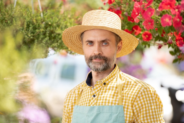 Mature gardener in hat and apron standing among flowers in bloom while working in greenhouse