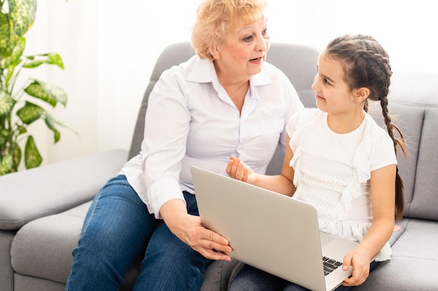 Mature grandmother helping child with homework at home. Satisfied old grandma helping her granddaughter studying in living room. Little girl writing on notebook with senior teacher sitting next to her