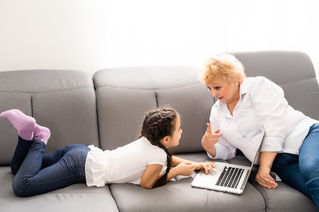 Mature grandmother helping child with homework at home. Satisfied old grandma helping her granddaughter studying in living room. Little girl writing on notebook with senior teacher sitting next to her