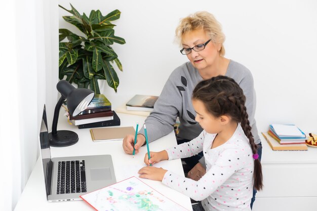 Nonna matura che aiuta il bambino a fare i compiti a casa. vecchia nonna soddisfatta che aiuta sua nipote a studiare in soggiorno. bambina che scrive sul taccuino con l'insegnante senior seduto accanto a lei